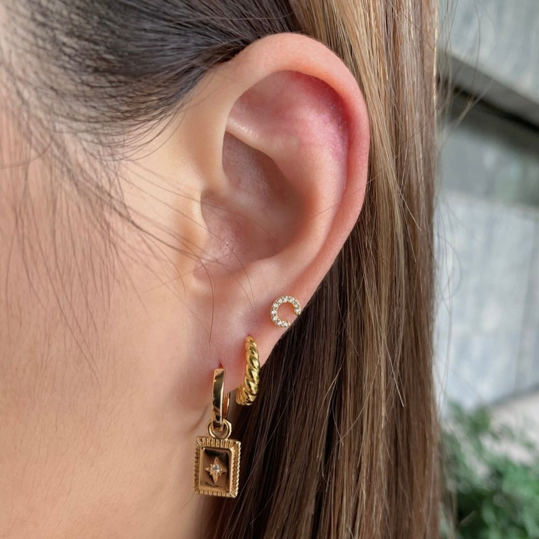 Charm Star Tag Huggies, Evocative of captivating talisman-inspired motifs, these gold hoop earrings are enhanced by an alluring charm centered with a dazzling cubic zirconia stone. Materials: Eco Brass 5A Cubic Zirconia 18K Gold Plating