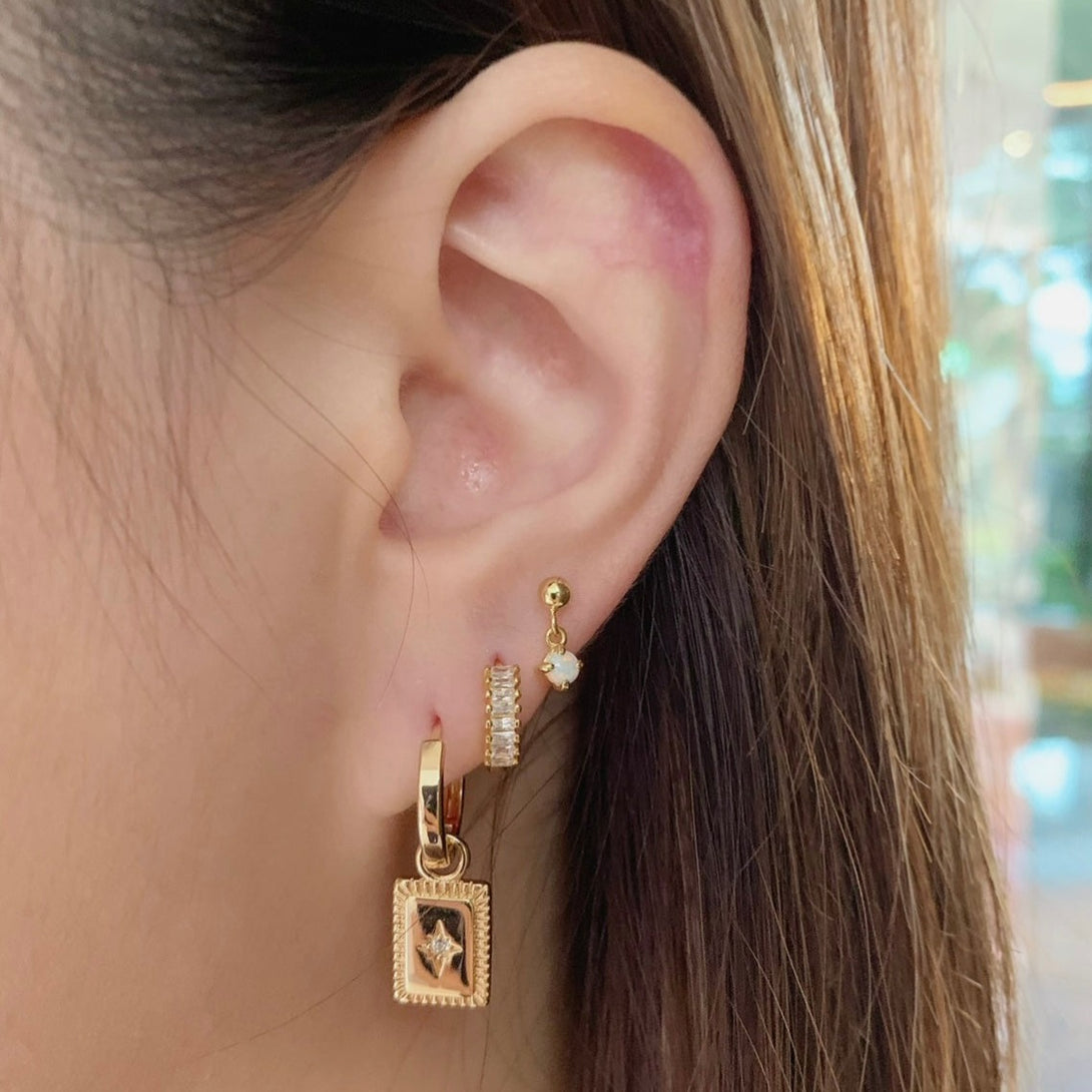 Charm Star Tag Huggies, Evocative of captivating talisman-inspired motifs, these gold hoop earrings are enhanced by an alluring charm centered with a dazzling cubic zirconia stone. Materials: Eco Brass 5A Cubic Zirconia 18K Gold Plating